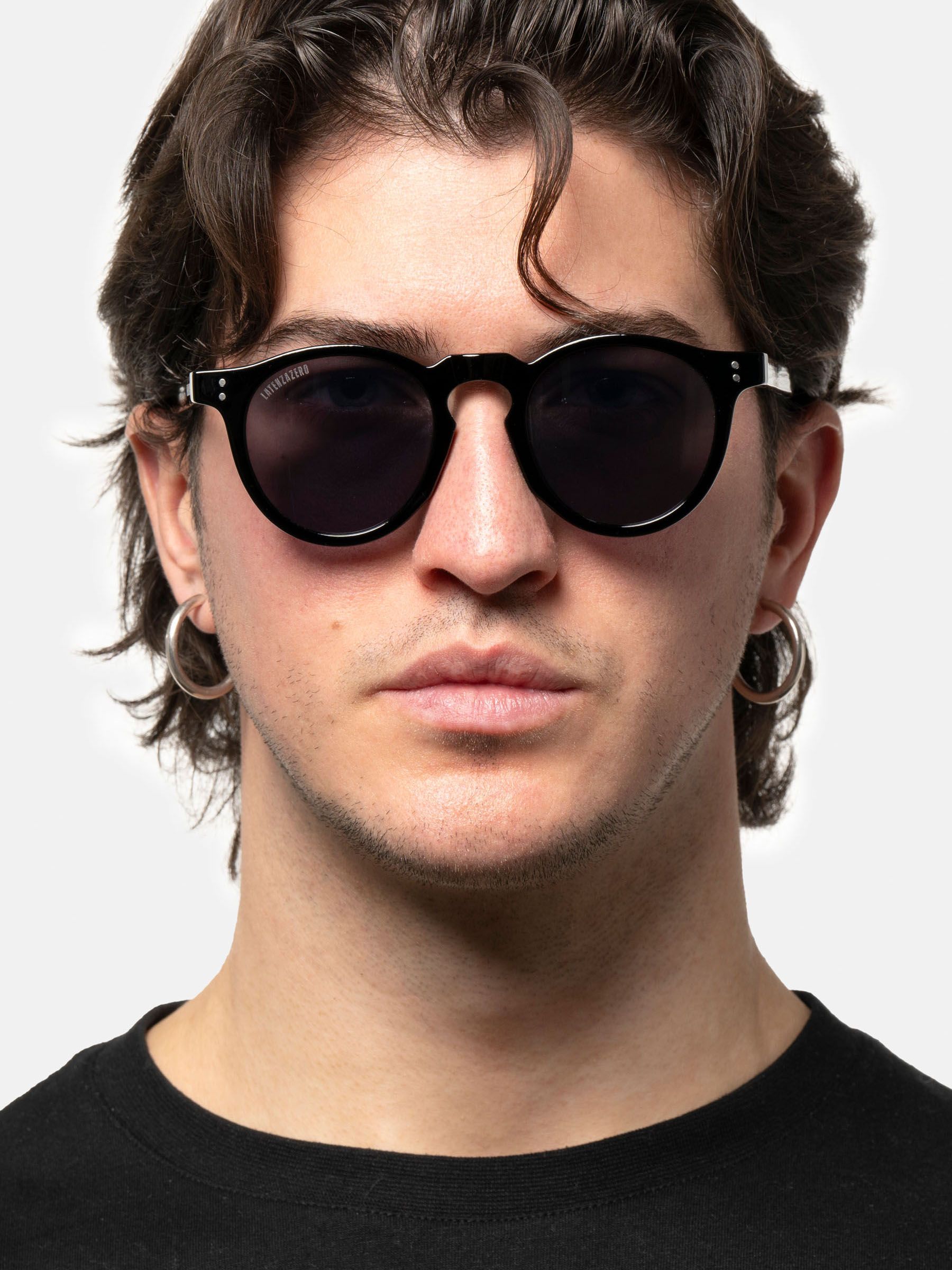 Un uomo indossa occhiali da sole neri con lenti rotonde. Gli occhiali sono sul suo viso. Indossa orecchini.