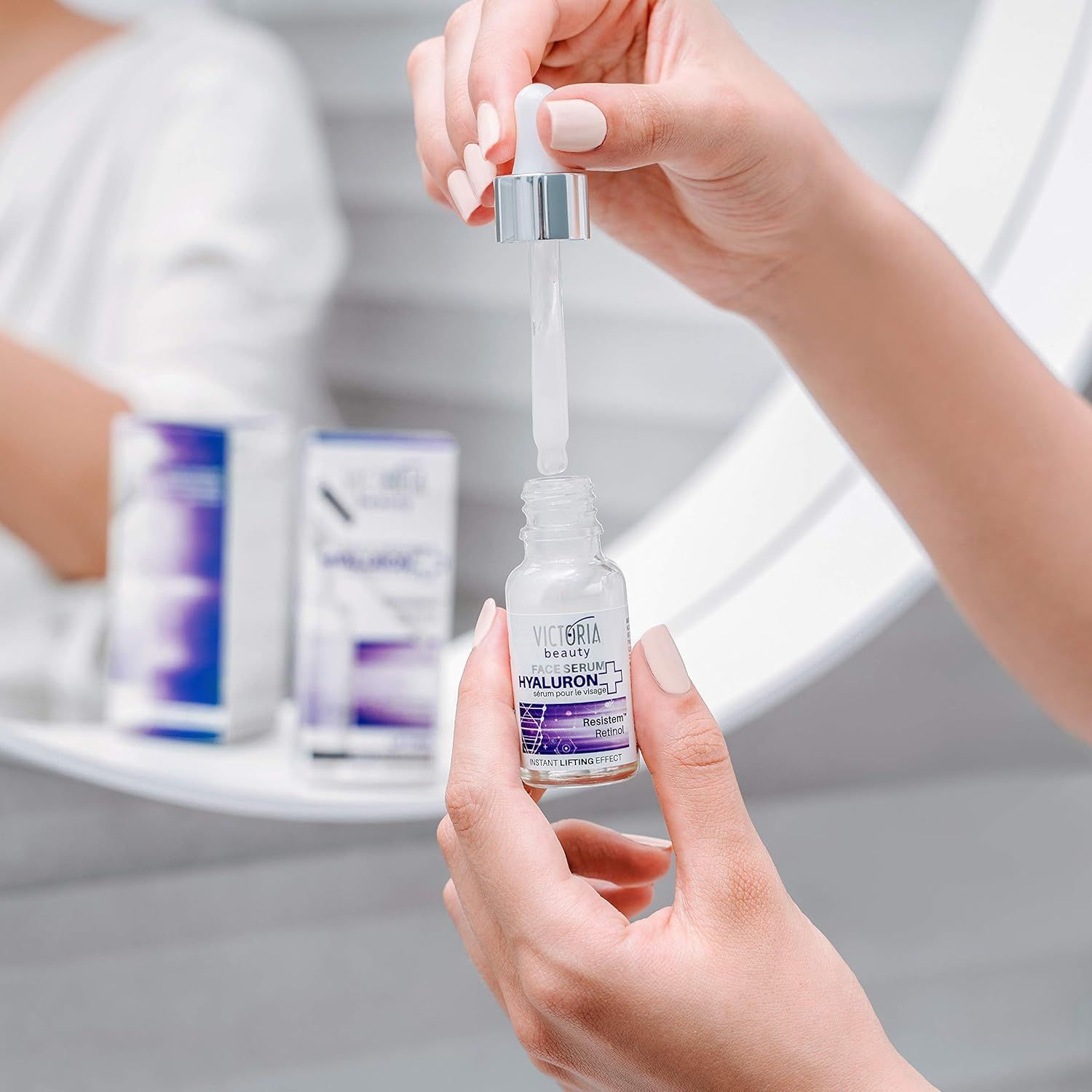 Mani che tengono un flacone di siero con pipetta. Sul flacone si legge "Victoria Beauty Hyaluron+". Un altro flacone e una confezione sullo sfondo.