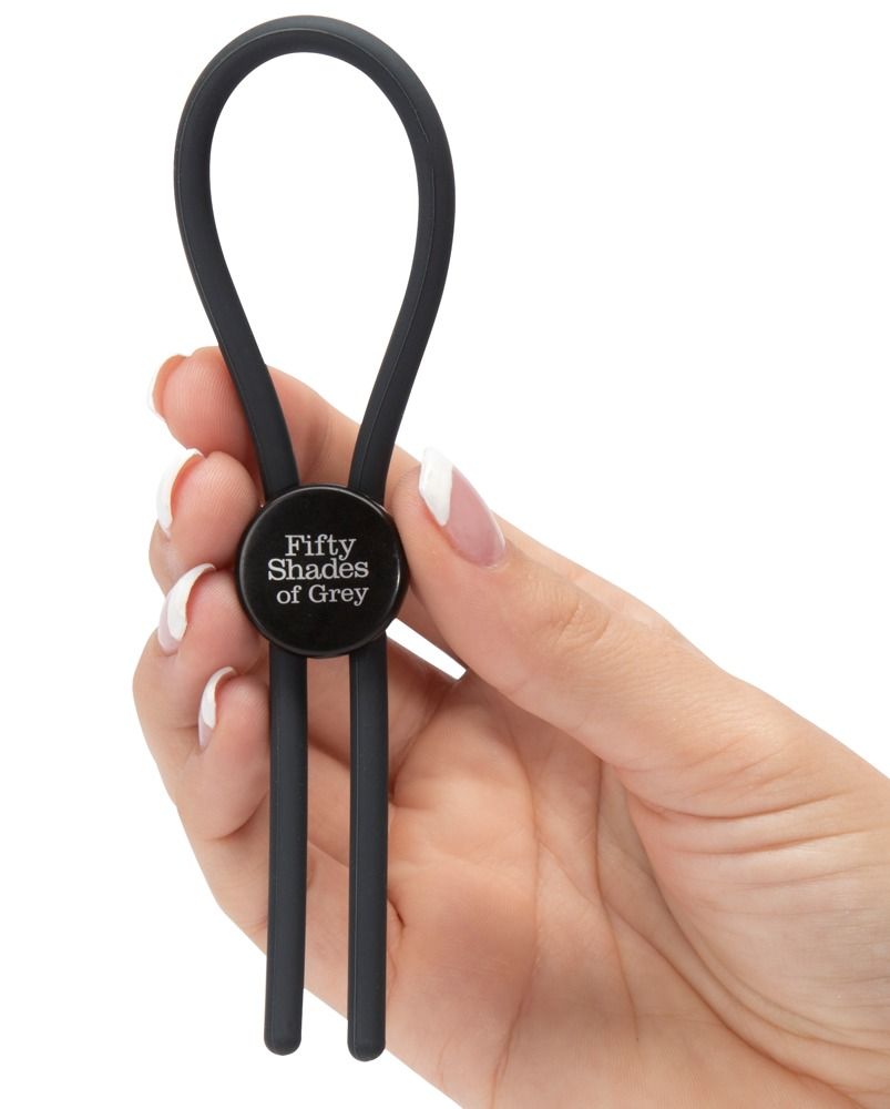 Anello fallico nero con logo Fifty Shades of Grey, tenuto in una mano. Due fili scendono.