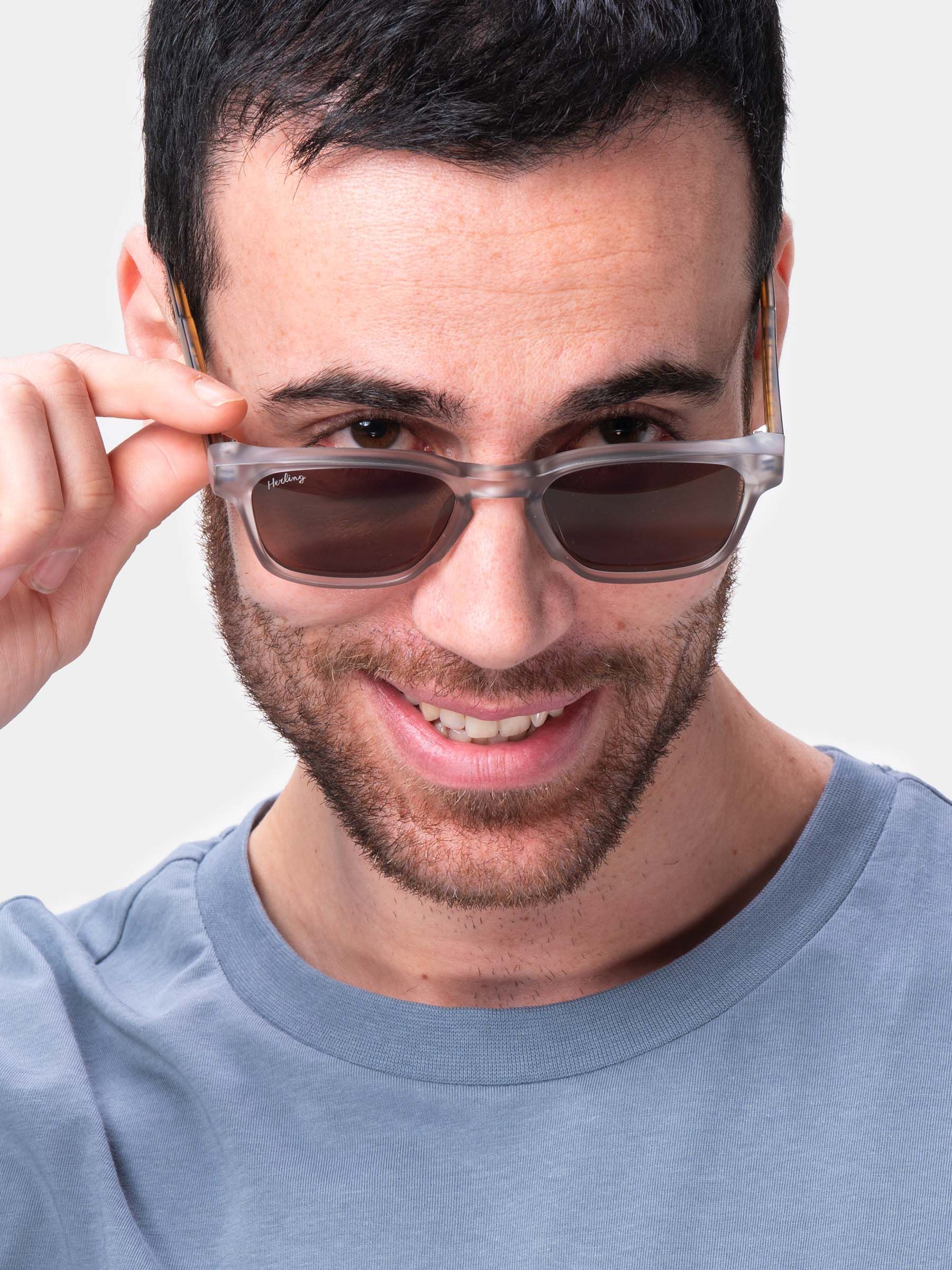 Uomo tiene occhiali da sole con lenti marroni e montatura grigia. Logo Herling sull'asta.