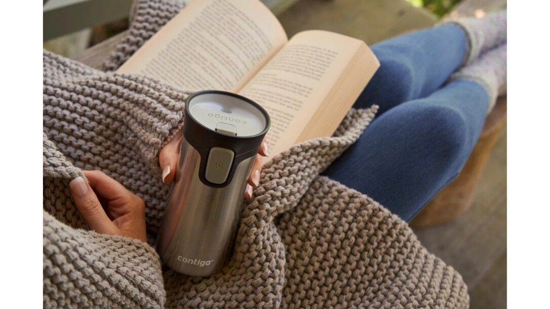 Persona che tiene un bicchiere termico. Bicchiere in acciaio inox, coperchio nero. Persona seduta con libro e coperta. Logo "Contigo" visibile.