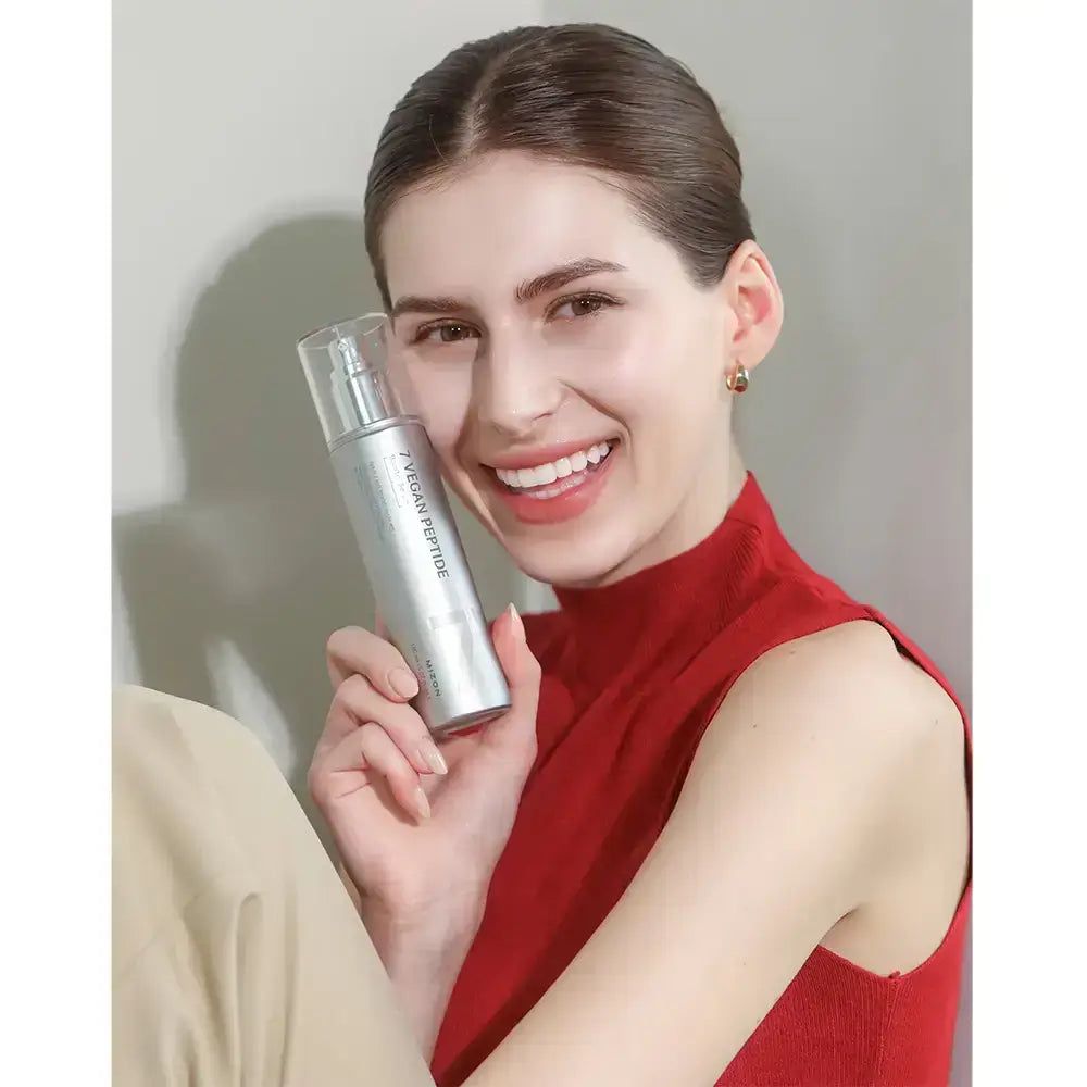 Donna tiene in mano una bottiglia d'argento. Sulla bottiglia c'è scritto "7 Vegan Peptide". La donna sorride. Indossa un top rosso e pantaloni beige.