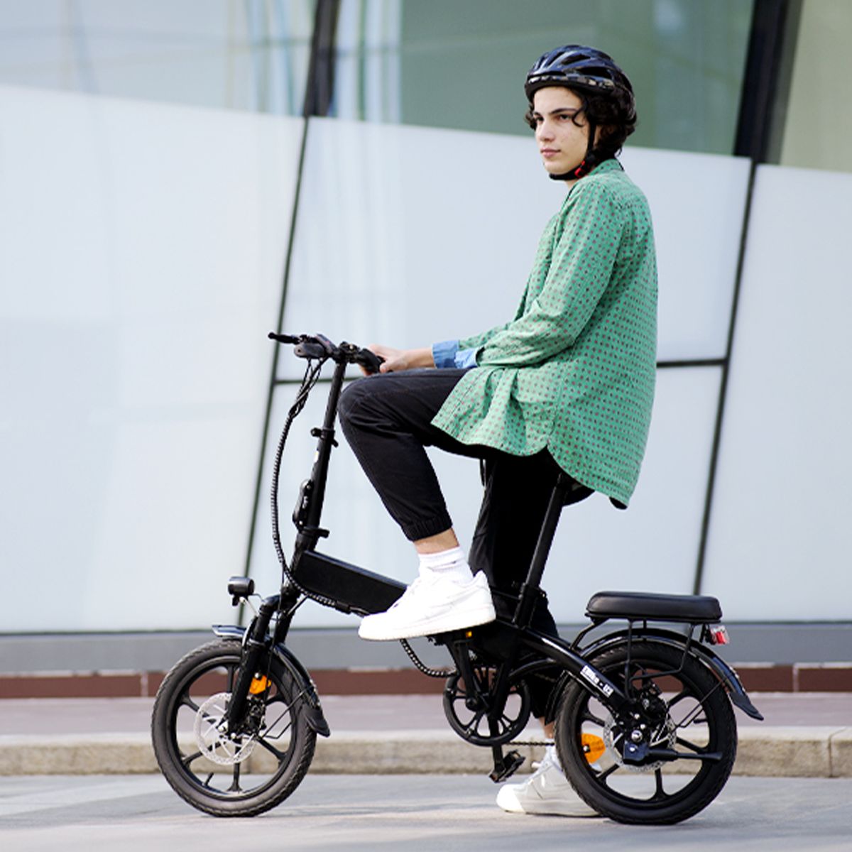 Persona seduta su una bicicletta elettrica pieghevole nera. Indossa casco e giacca verde. Sfondo: edificio.