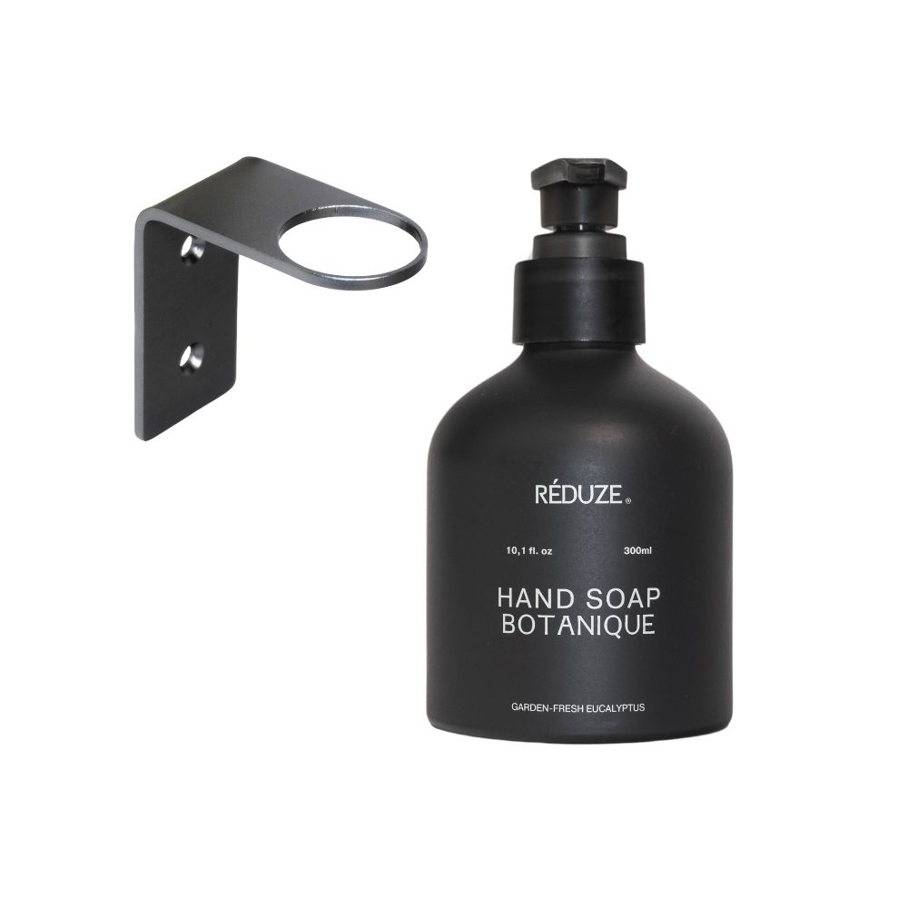 Sapone per le mani nero con supporto a parete. Scritta: HAND SOAP BOTANIQUE. Erogatore nero con testa a pompa. Supporto a parete in metallo.