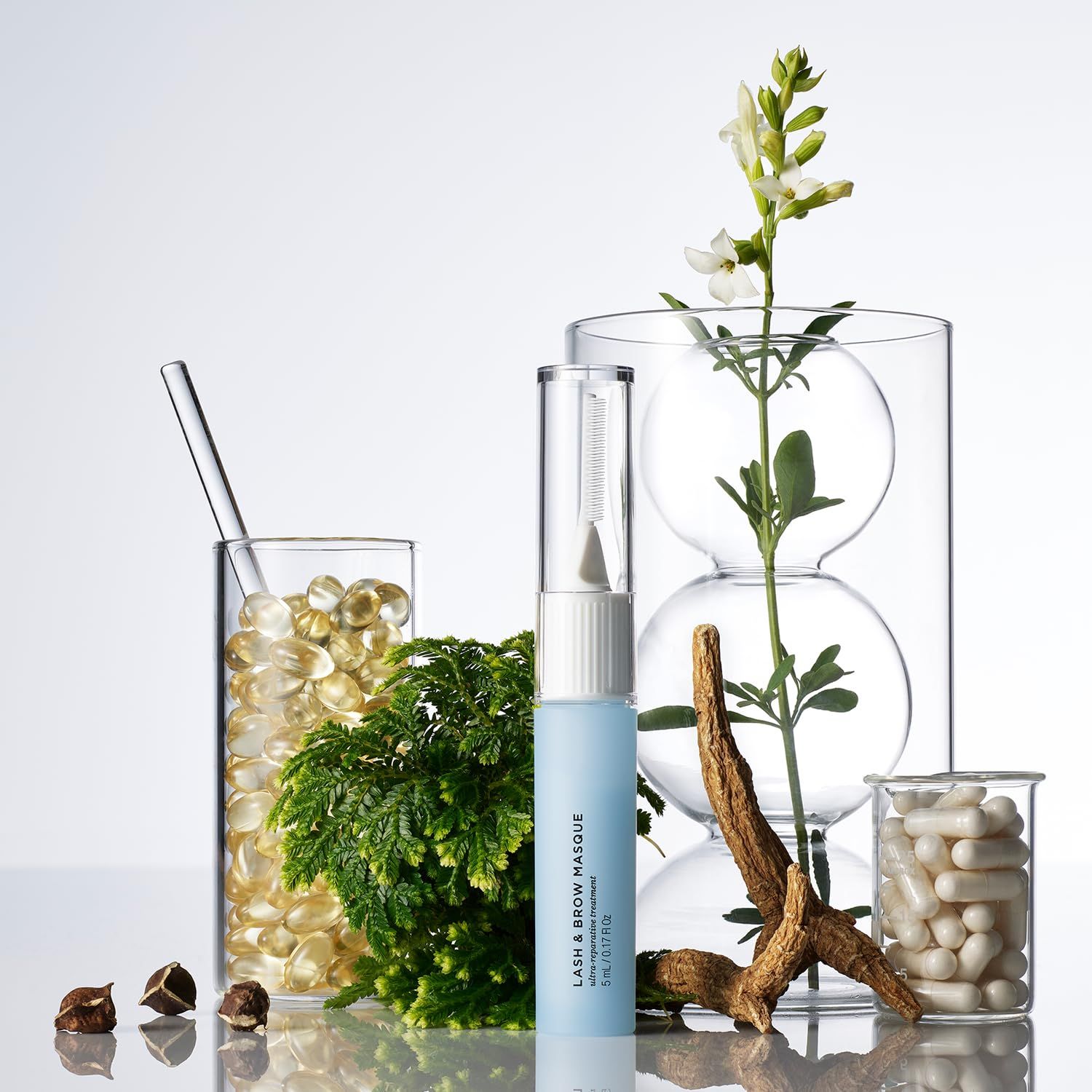 Flacone azzurro con applicatore bianco, circondato da piante e contenitori. Testo: LASH & BROW MASQUE.