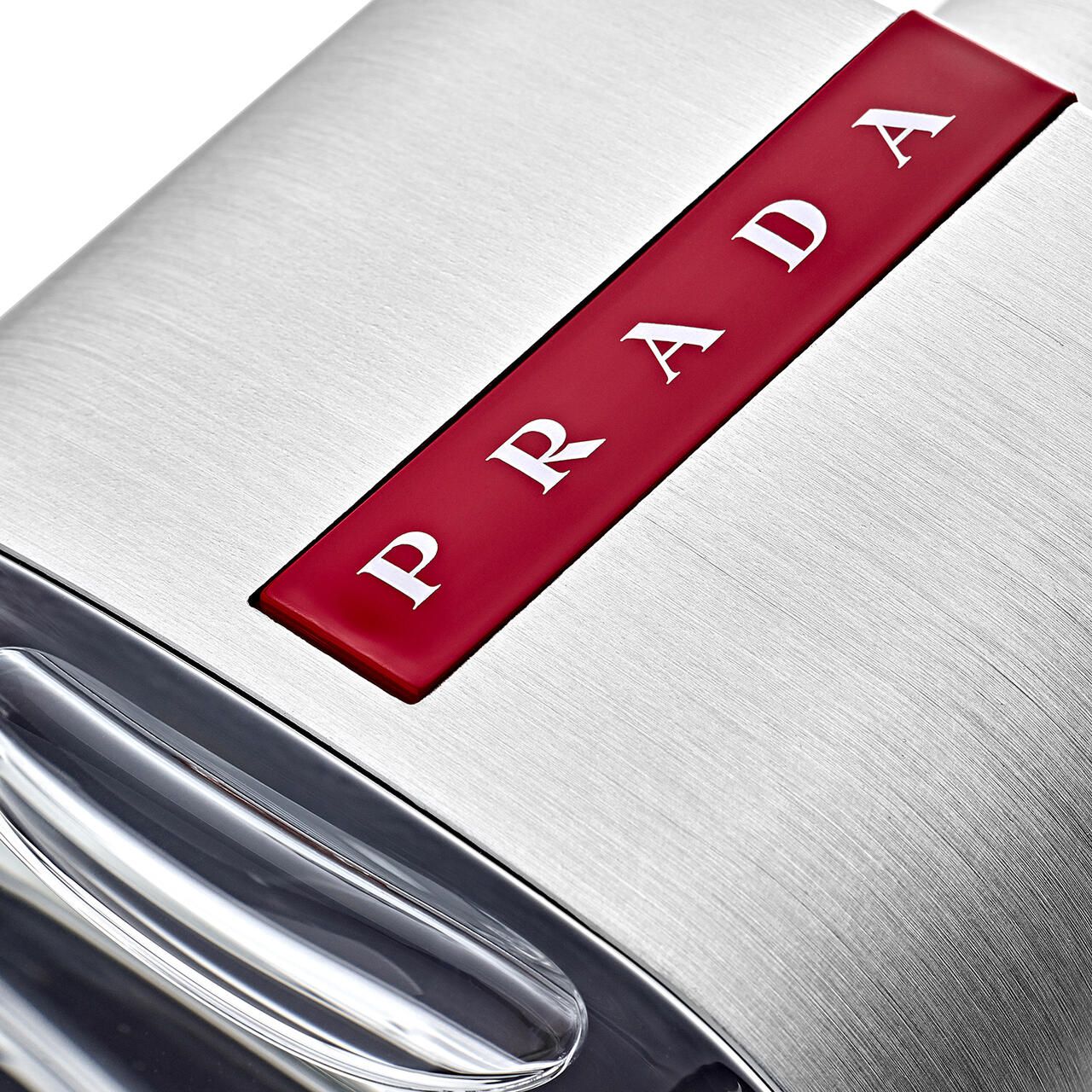 Primo piano del logo Prada su un flacone argentato. Striscia rossa con scritta Prada bianca.