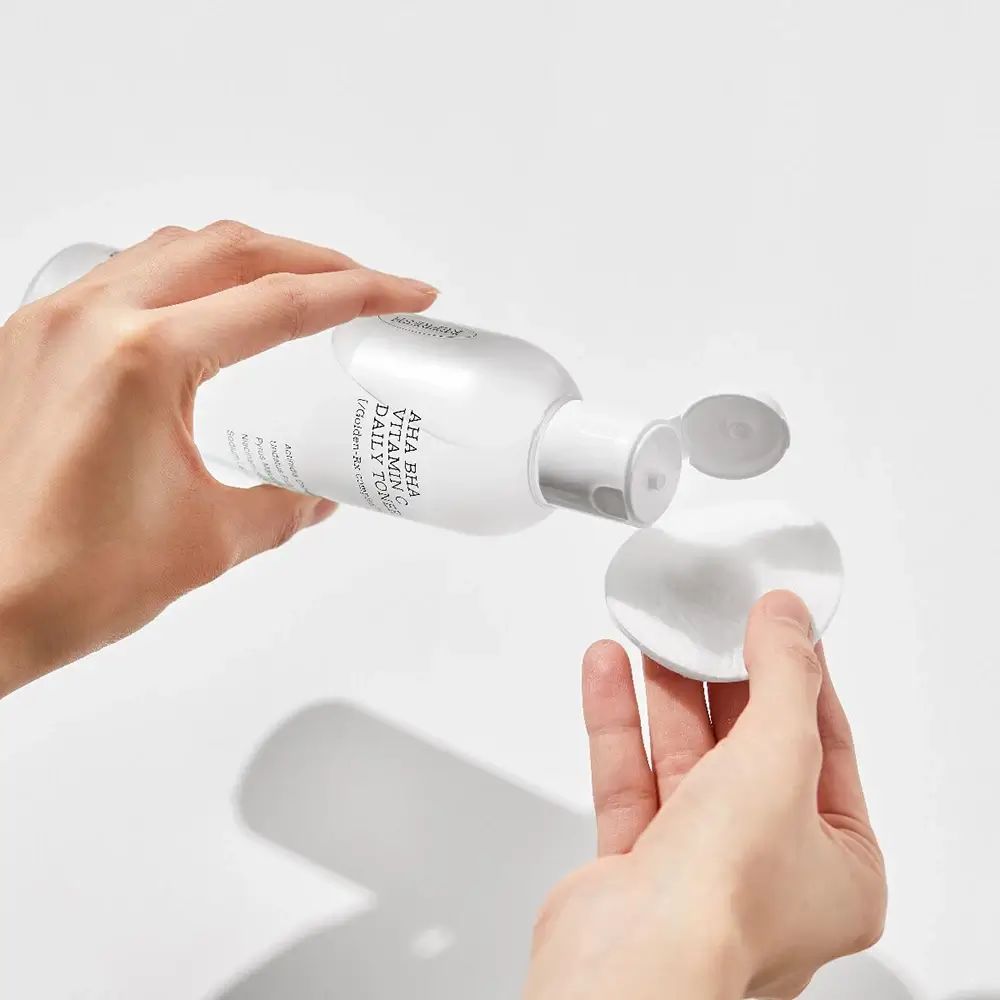Flacone bianco di toner versato su un dischetto di cotone. Scritta: AHA BHA Vitamin C Daily Toner.