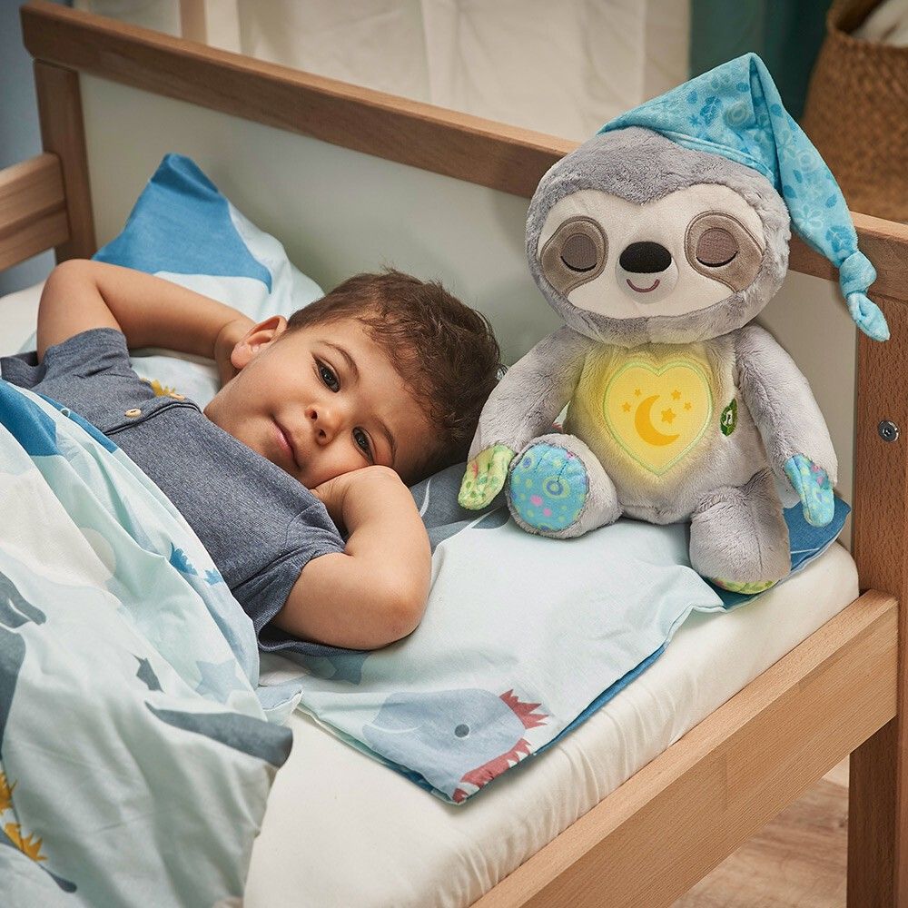 Bambino a letto accanto a un peluche bradipo grigio con cappello. Cuore giallo con motivo luna-stella.