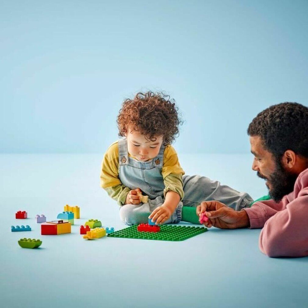 Bambino e adulto giocano con mattoncini LEGO Duplo su una base verde. Sfondo blu.