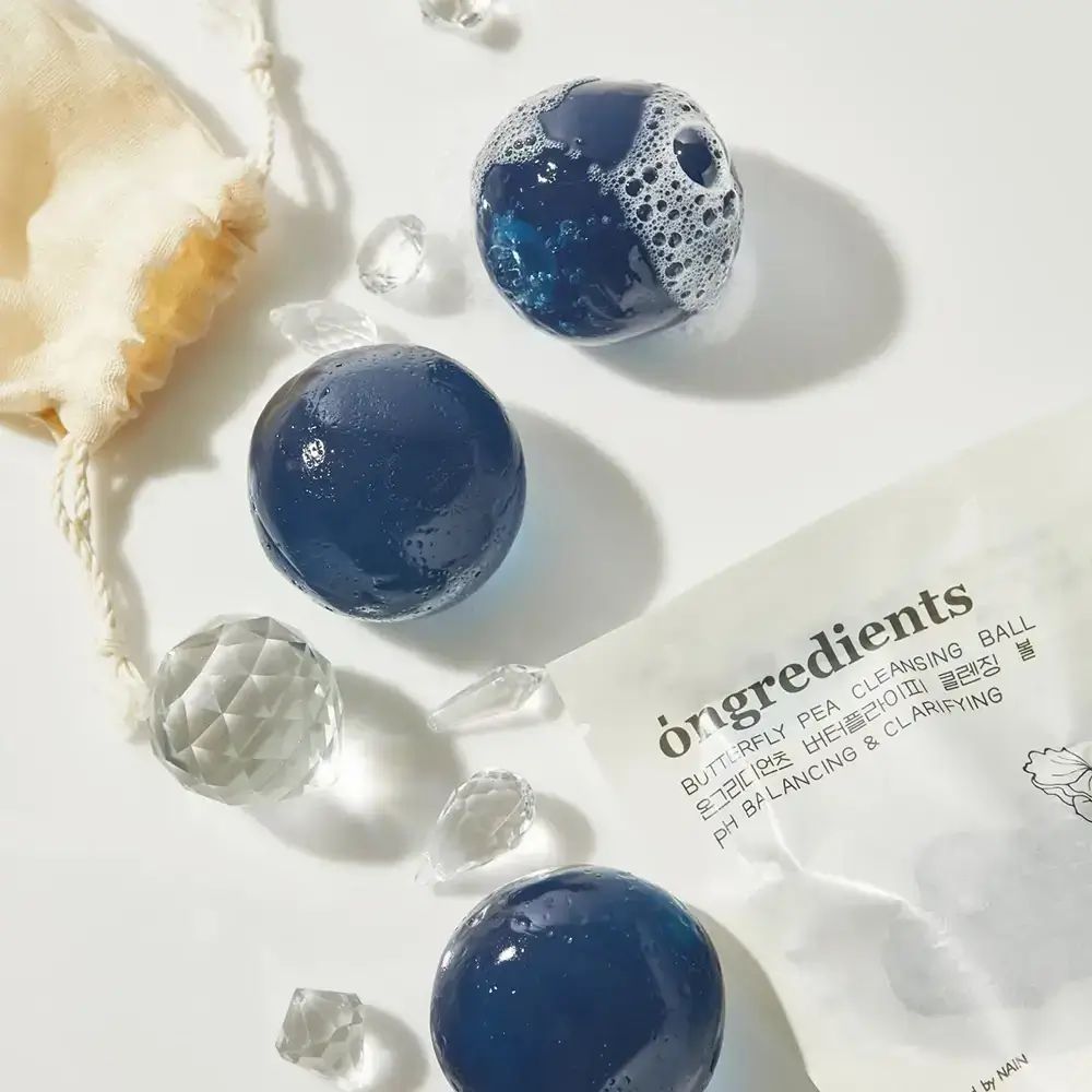 Tre sfere blu, una con schiuma, accanto a confezione e cristalli. Su sfondo bianco.