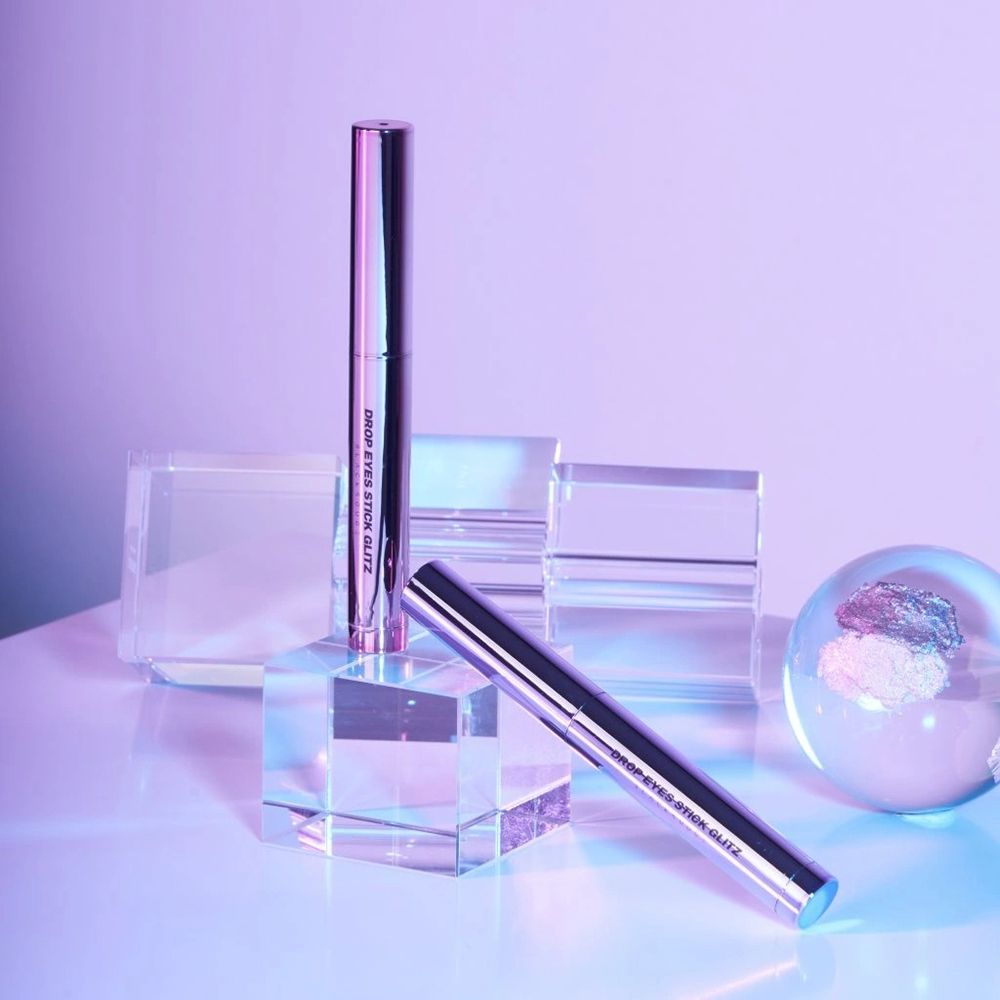 Due Drop Eyes Stick Glitz Eyeshadow metallici. Un bastoncino in piedi, uno sdraiato. Posizionati su blocchi trasparenti e una sfera.