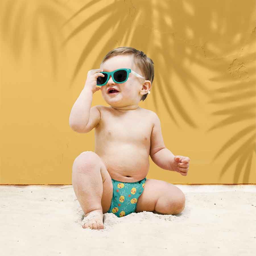 Bambino con occhiali da sole e costume da bagno con ananas sulla spiaggia. Sfondo giallo con ombre.