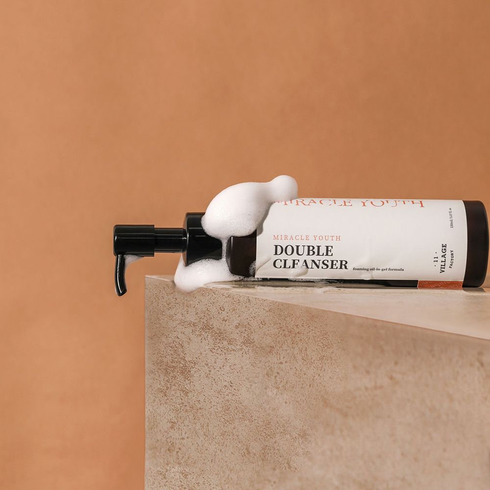 Flacone marrone con dosatore nero, schiuma che esce. Scritta: MIRACLE YOUTH, DOUBLE CLEANSER. Su una superficie.