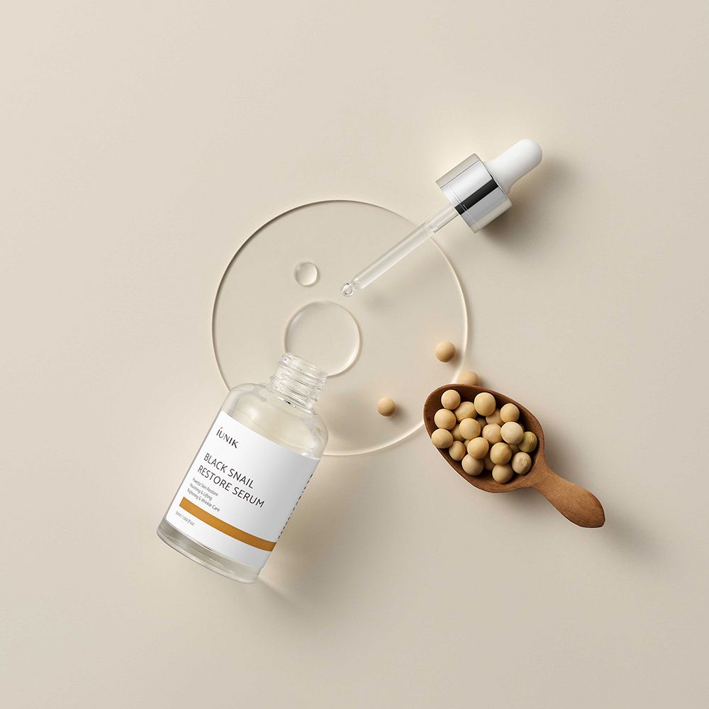 Flacone aperto con pipetta, accanto a paletta di legno con fagioli e disco trasparente. Prodotto: iUNIK Black Snail Restore Serum.