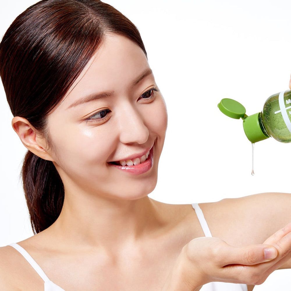 Donna con pelle luminosa che versa olio detergente da una bottiglia verde nella sua mano.