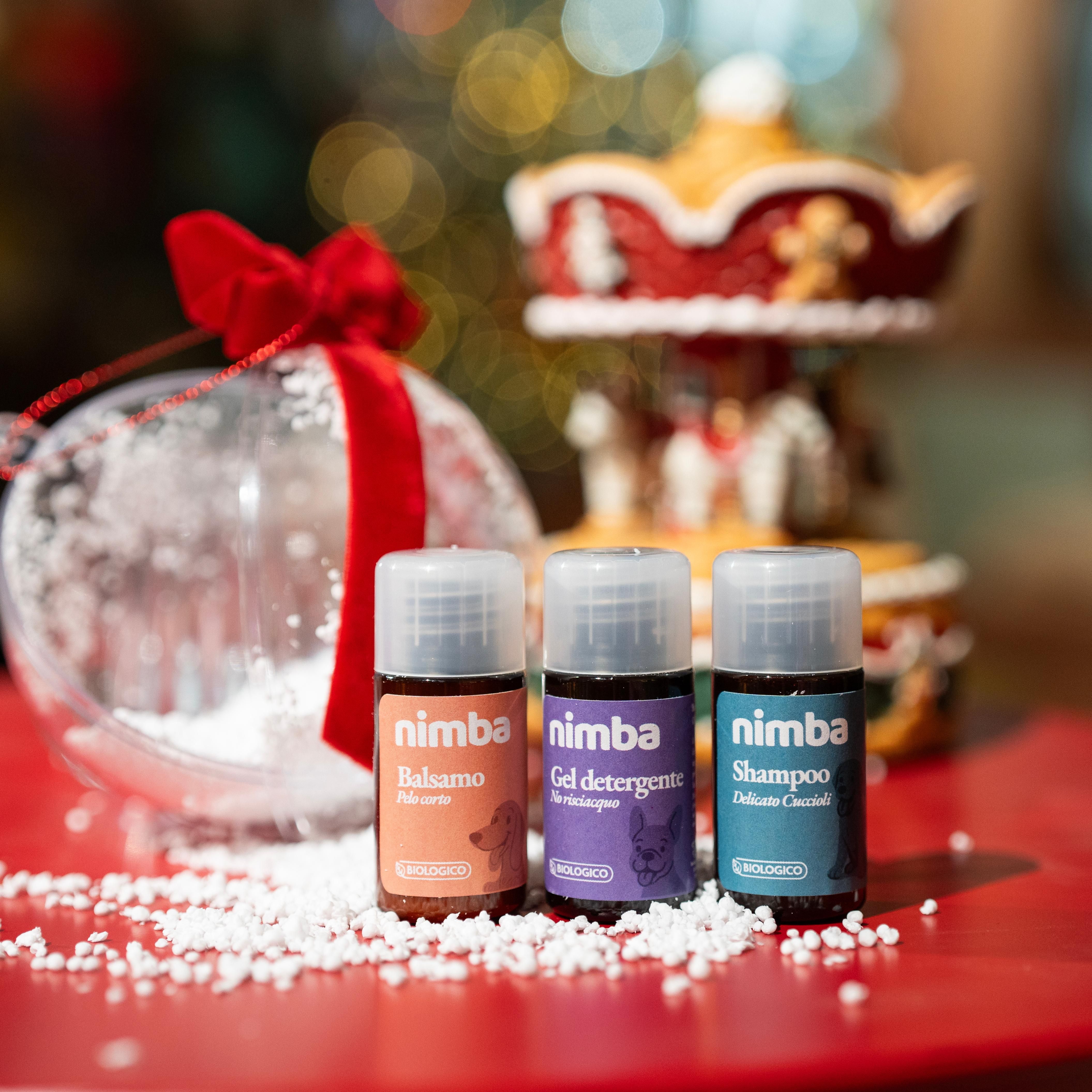 Tre piccoli flaconi "nimba" nella neve finta su tavolo rosso. Decorazioni natalizie sullo sfondo.