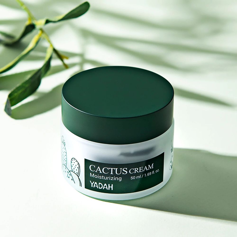 Barattolo di crema con coperchio verde. Scritta: Cactus Cream, Yadah. 50 ml. Illustrazione di cactus. Sfondo con foglie.