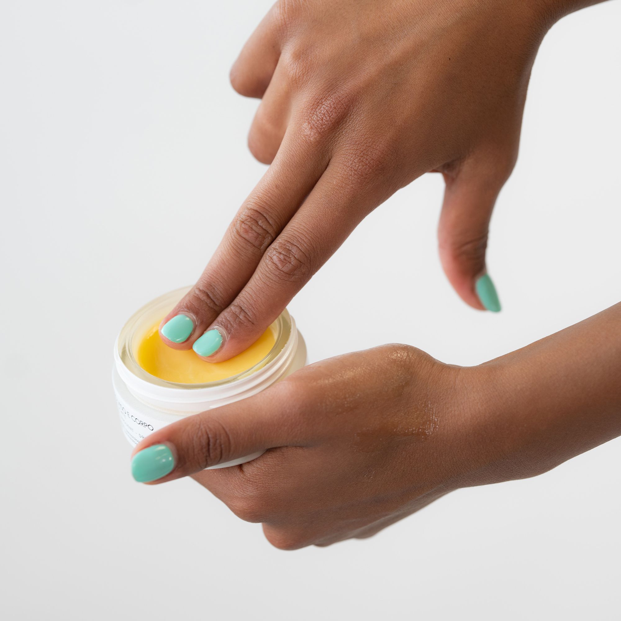 Mani che toccano la crema in un vasetto bianco. Crema gialla visibile. Pelle chiara, unghie turchesi.