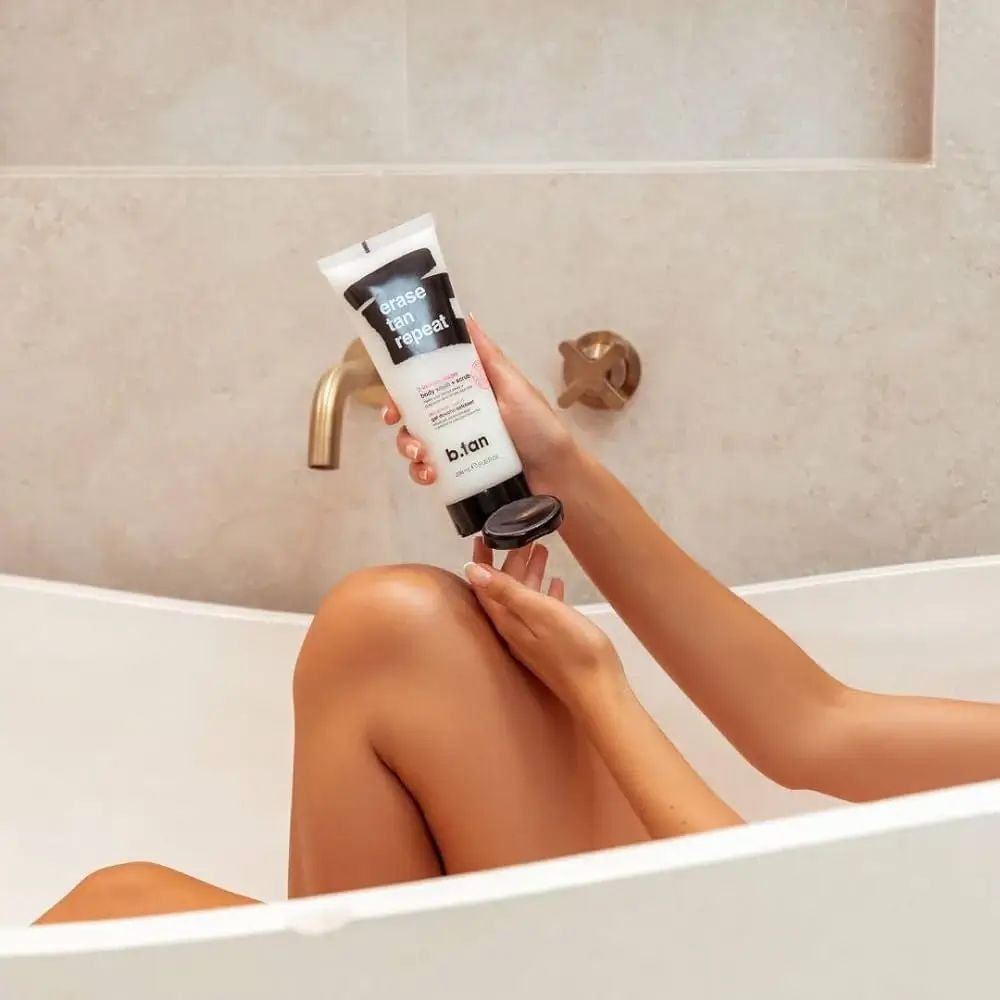 Persona tiene un tubetto sopra una gamba in una vasca da bagno. Sul tubetto c'è scritto "erase tan repeat" e "b.tan".
