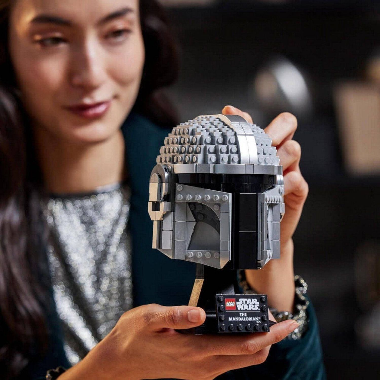 Donna tiene casco LEGO Star Wars. Casco grigio-argento su supporto. Loghi LEGO, Star Wars. Ambiente luminoso.