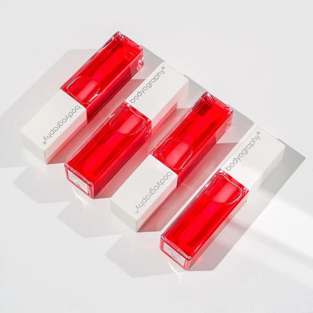 Quattro flaconi di olio per labbra con coperchi bianchi. Il marchio Bodyography è stampato sui coperchi. I flaconi sono riempiti di rosso.