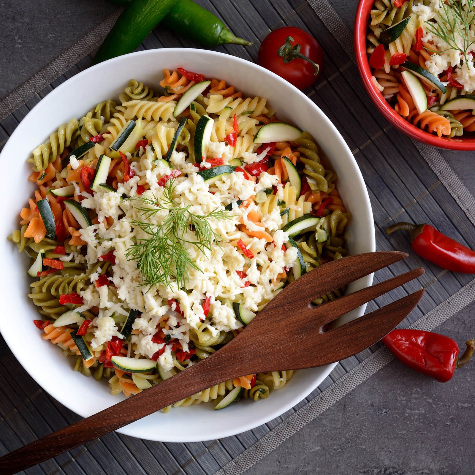 Ciotola bianca con fusilli tricolore, zucchine, peperoni e formaggio. Forchetta di legno e peperoni rossi accanto.
