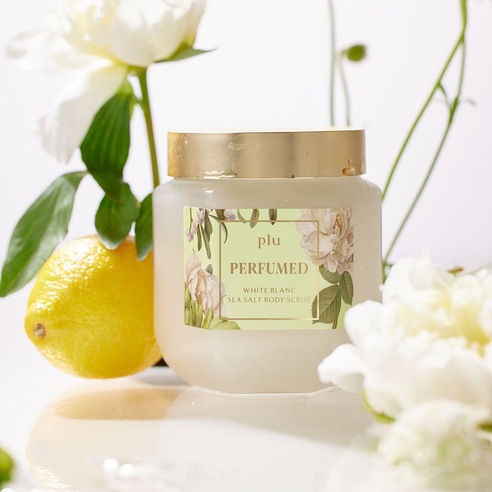 Barattolo di vetro con coperchio dorato ed etichetta. L'etichetta recita "PLU PERFUMED White Blanc Sea Salt Body Scrub". Limone e fiori bianchi.