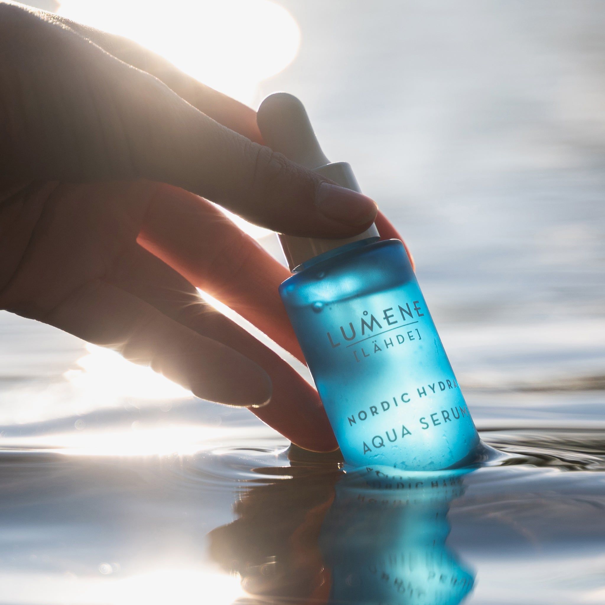 Flacone blu in acqua, tenuto da una mano. Scritta: Lumene [LAHDE] Nordic Hydra Aqua Serum.