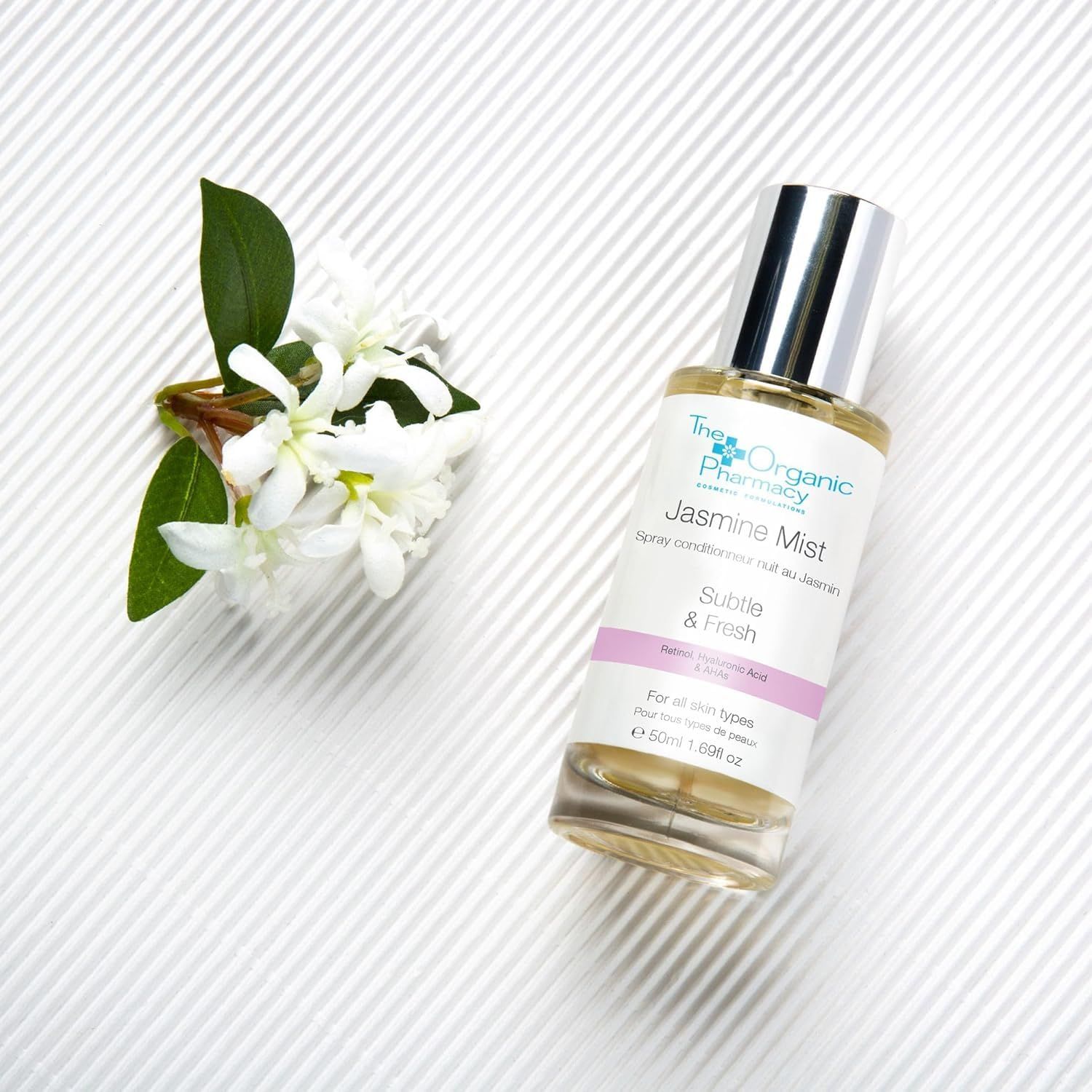 Flacone in vetro con tappo argentato e fiori su sfondo bianco. Etichetta rosa con testo. Marchio: The Organic Pharmacy.