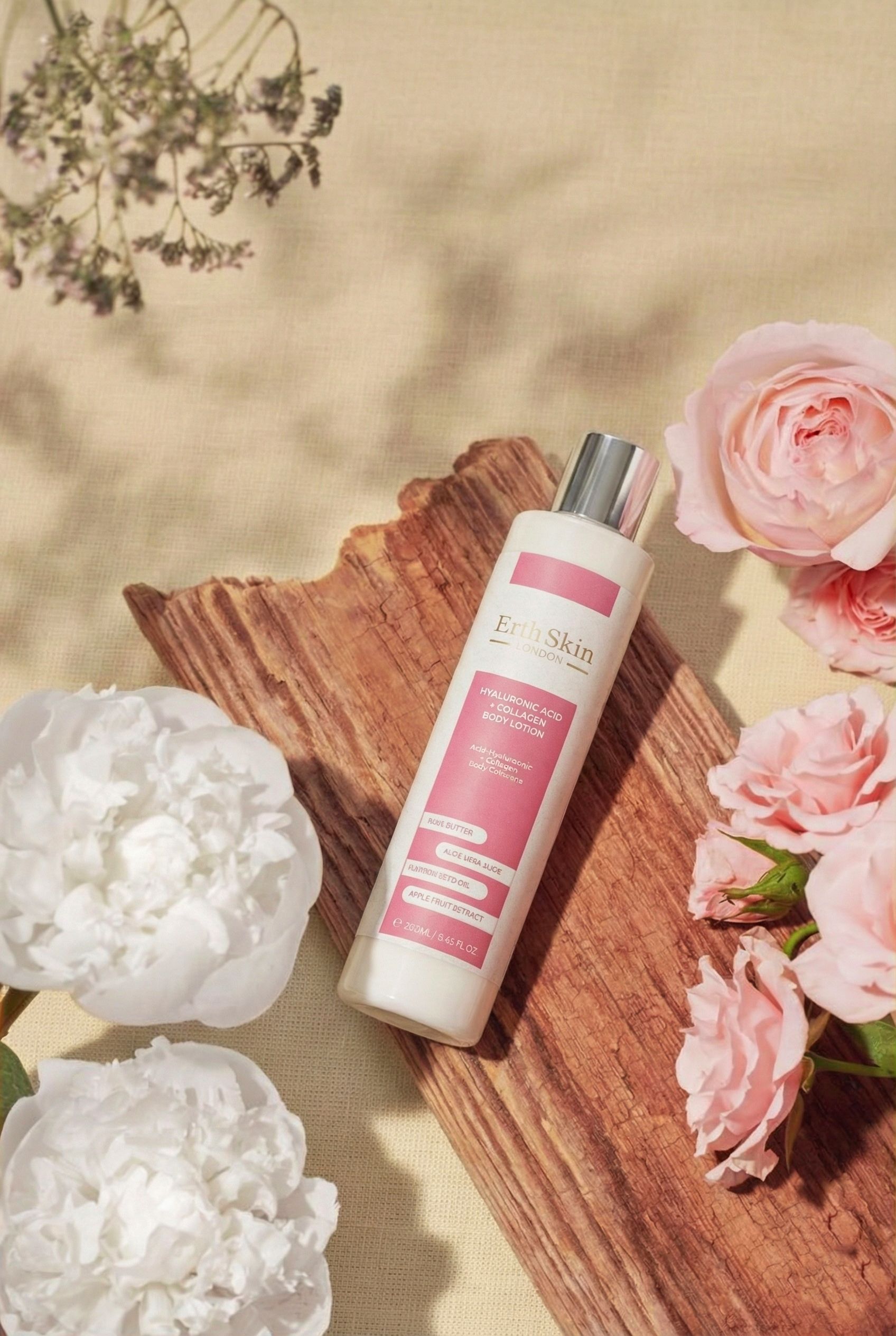 Flacone di lozione per il corpo bianco con etichetta rosa. Contiene acido ialuronico e collagene. Di ErthSkin London. Posizionato su legno e fiori.