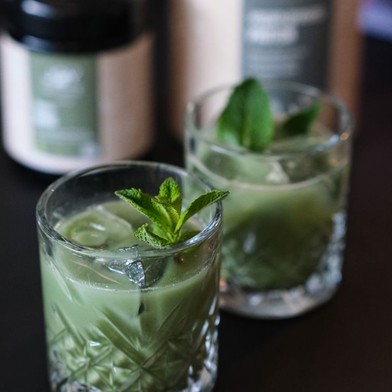 Due bicchieri con bevanda verde, cubetti di ghiaccio e menta. Altri prodotti sullo sfondo. Tavolo nero.