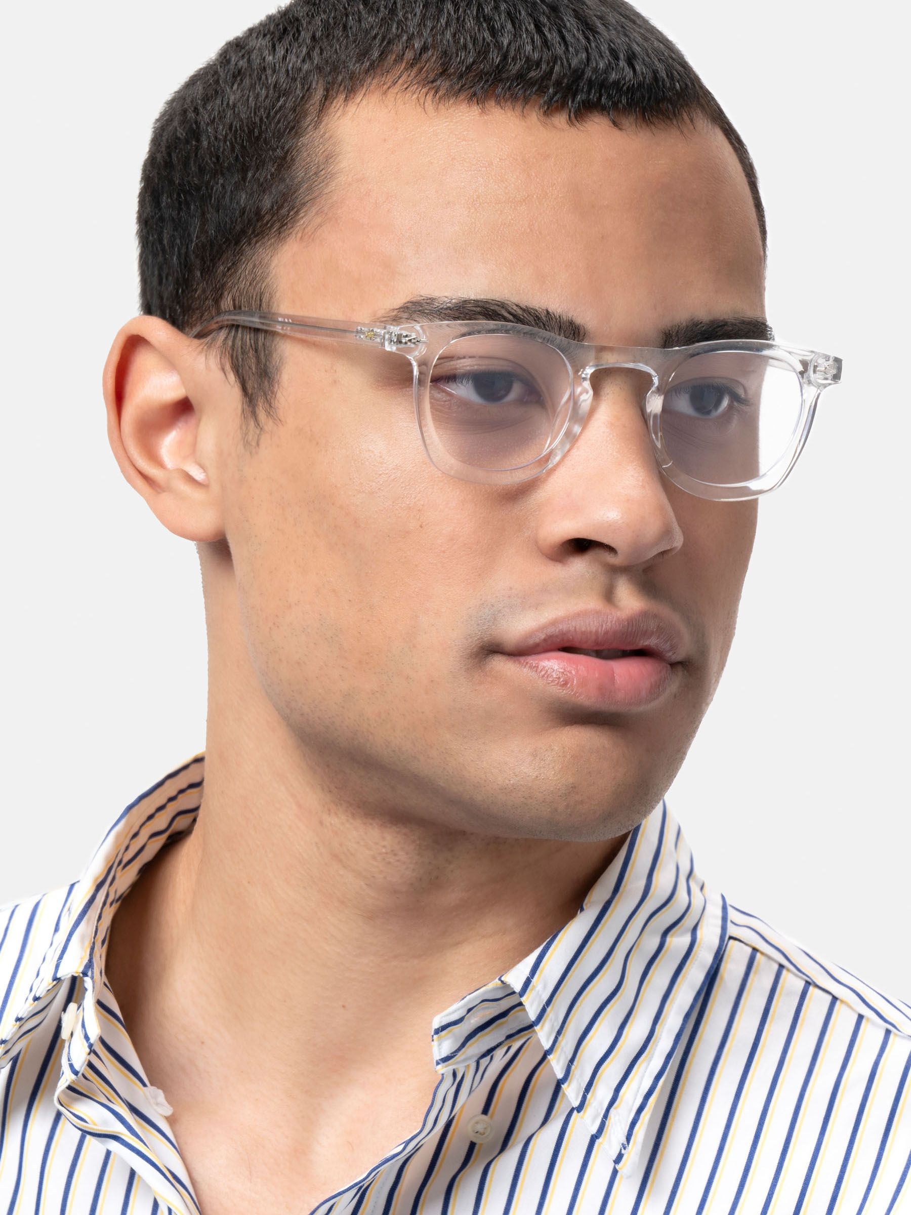 Uomo con occhiali. Montatura trasparente, lenti rotonde. Camicia a righe. Sguardo laterale.