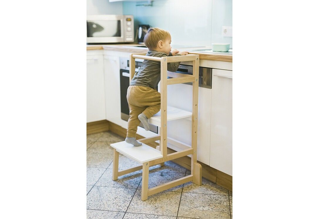 Bambino che sale su una torre di apprendimento in una cucina. Struttura in legno con gradini bianchi. Il bambino afferra un oggetto sul piano di lavoro.