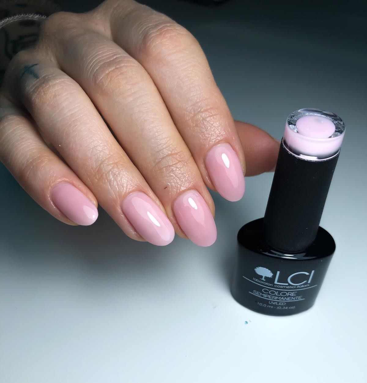Mano con unghie smaltate di rosa e flacone di smalto. Flacone con logo e testo. Unghie di forma ovale, lucide.