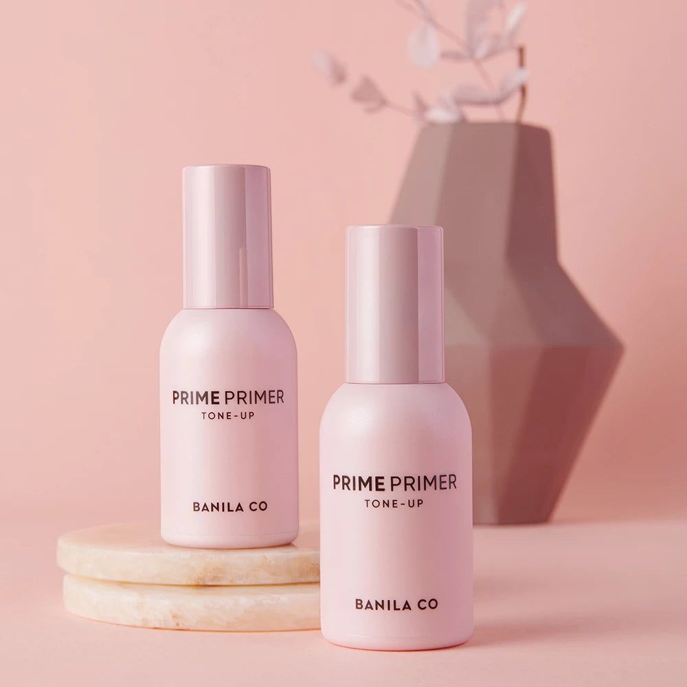 Due flaconi di primer rosa con tappi. Scritta: PRIME PRIMER TONE-UP, BANILA CO. Su un piedistallo.