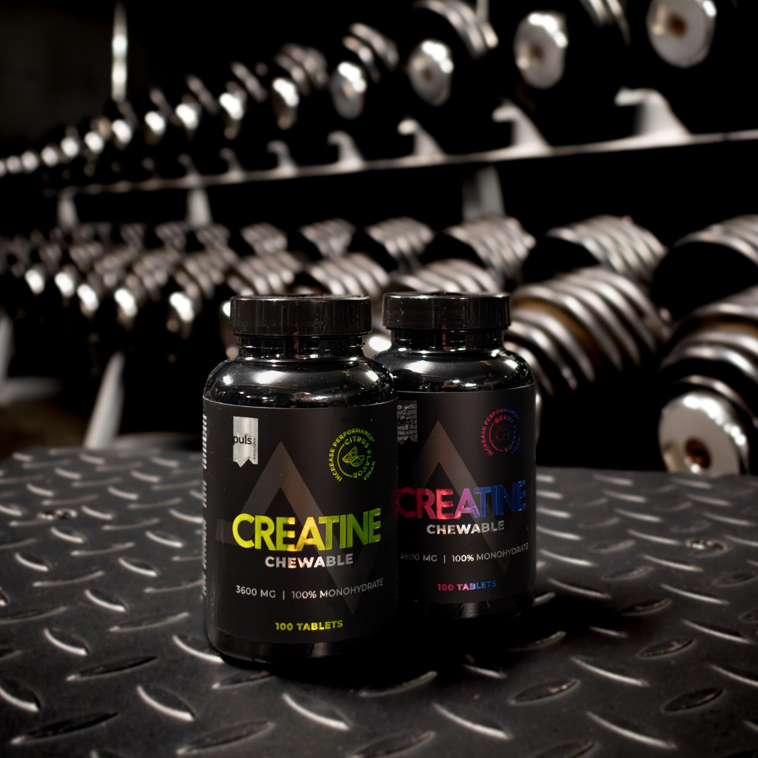 Due barattoli neri "Creatine Chewable" davanti a uno scaffale per manubri in palestra.