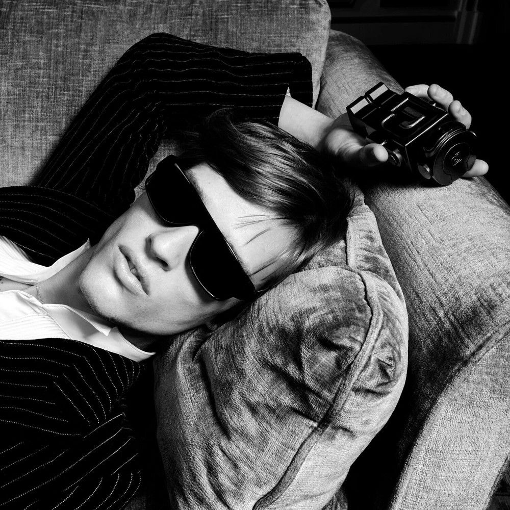 Foto in bianco e nero di un uomo in abito e occhiali da sole che si riposa su un divano, con un flacone di profumo.