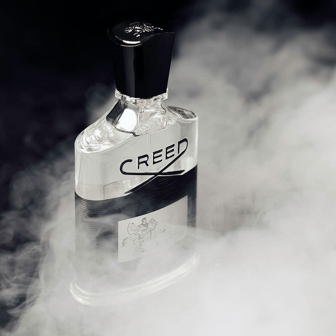 Flacone in vetro con tappo nero. Scritta "CREED". Etichetta nera con "AVENTUS". Sfondo nebbioso.