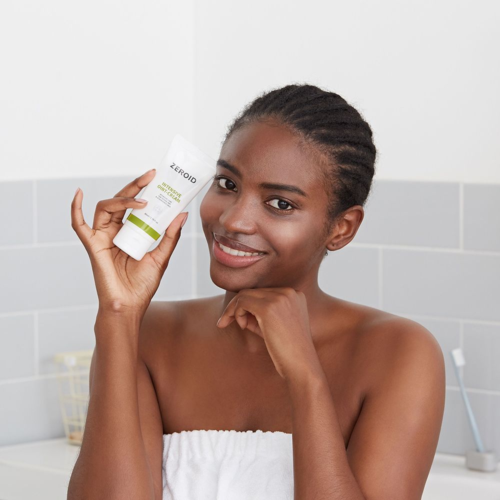 Donna tiene tubo ZEROID INTENSIVE OINT-CREAM. Sorride. Sfondo: bagno con piastrelle.