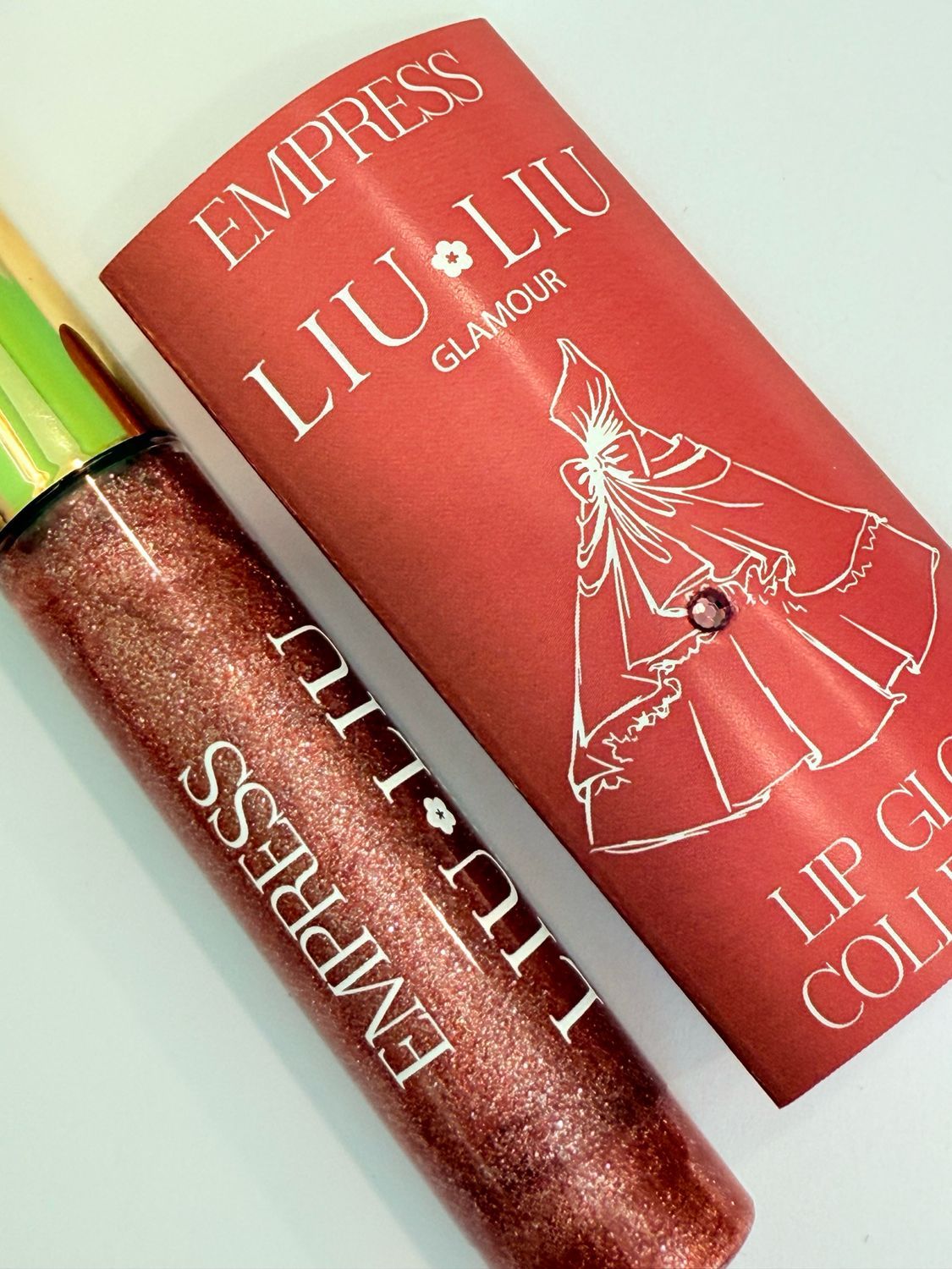 Flacone di lucidalabbra e confezione. Scritta: EMPRESS LIU LIU Glamour. Lucidalabbra rosso con tappo dorato.