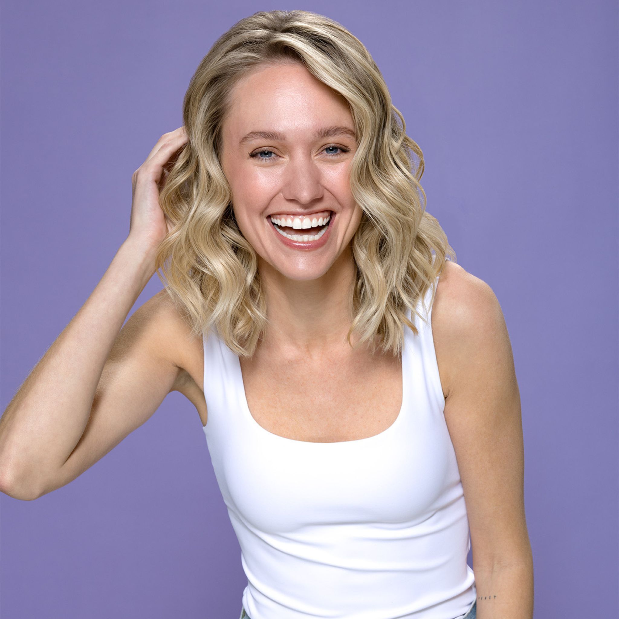 Donna sorridente con capelli biondi. Indossa una canotta bianca. Sfondo viola.