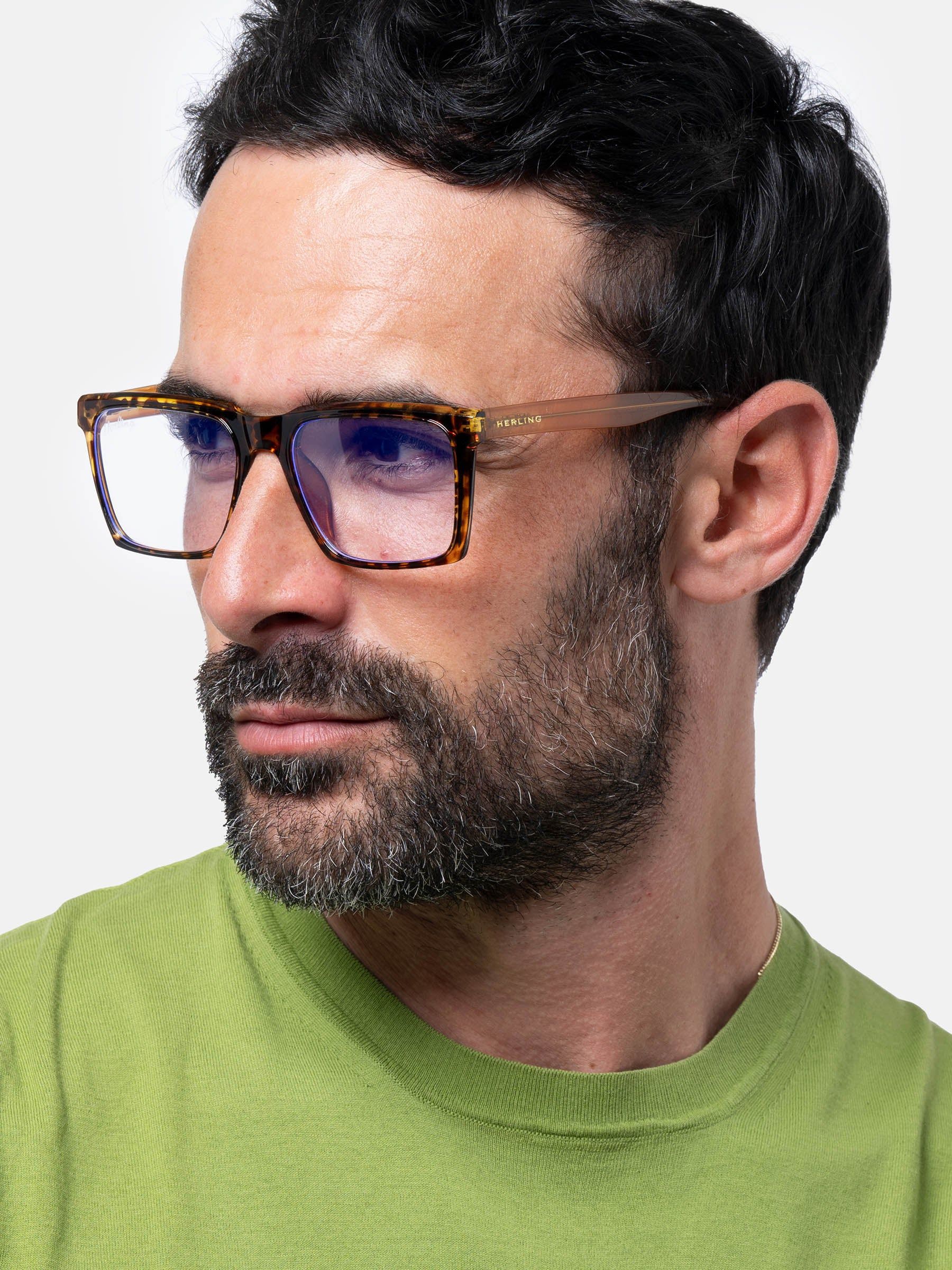 Uomo con occhiali. Occhiali con montatura marrone e lenti rettangolari. Aste e montatura con motivo tartarugato. Logo Herling.