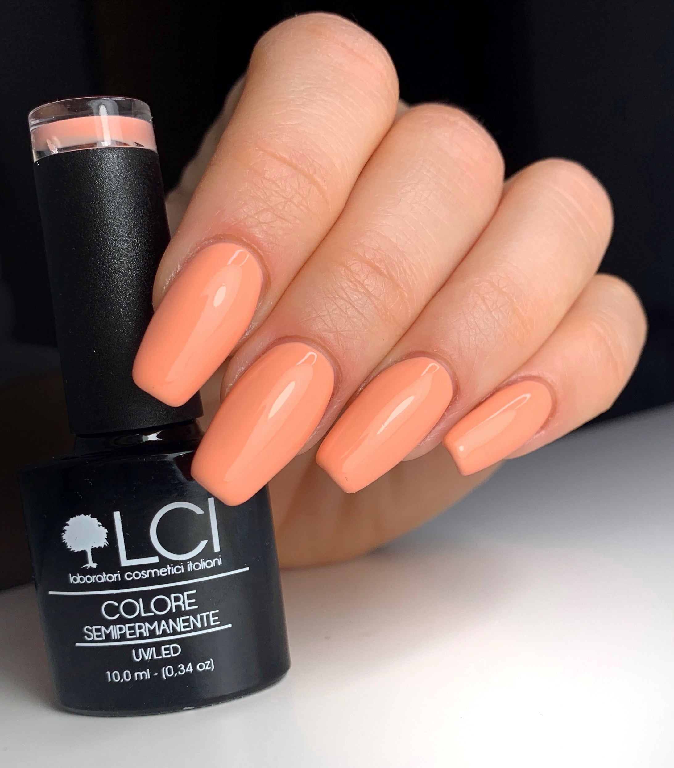 Mano con unghie smaltate e flacone di smalto. Unghie rosa. Flacone nero. Testo: LCI, Colore Semipermanente.