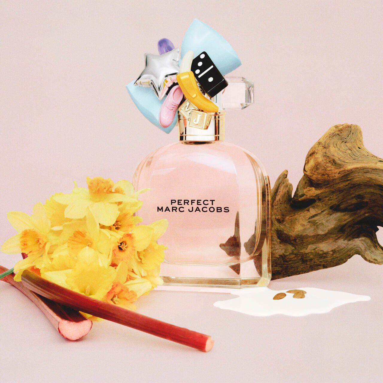 Flacone di profumo con tappo decorativo. Scritta: PERFECT MARC JACOBS. Accanto al flacone: fiori, pezzo di legno, liquido.