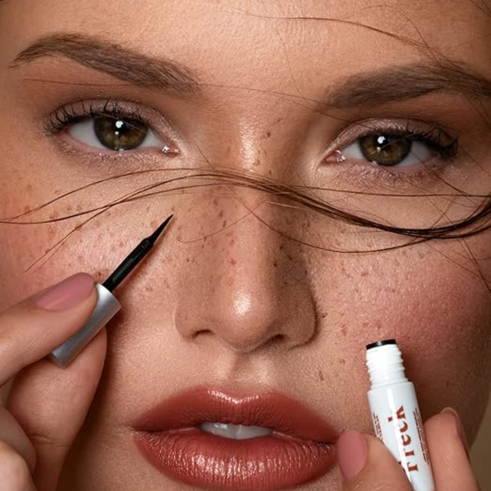 Persona tiene due flaconi. Uno con applicatore nero, l'altro con scritta "Freck". Viso con lentiggini.