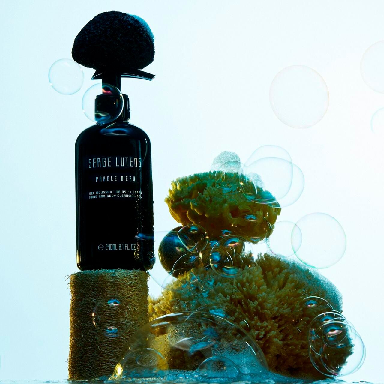 Flacone nero con dosatore su piedistallo. Testo: Serge Lutens, Parole d'eau, Gel m. et corps, Hand and body cleansing gel. Bolle di sapone.