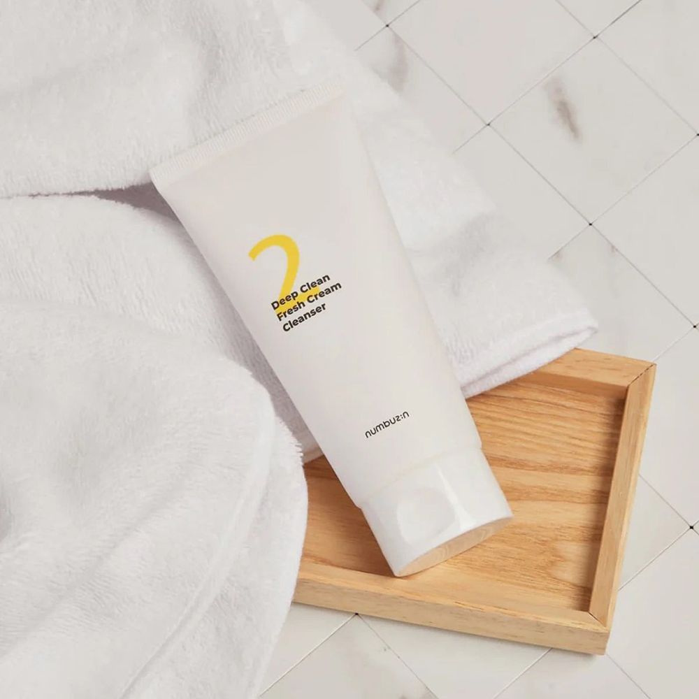Tubo bianco con "2" giallo e scritta "Deep Clean Fresh Cream Cleanser" su vassoio di legno e asciugamano.