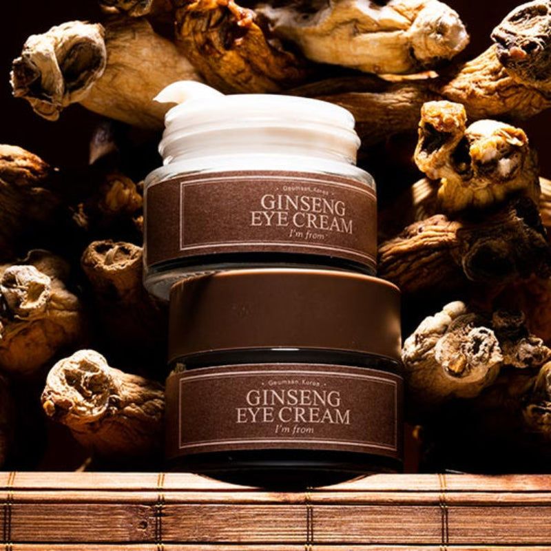 Due vasetti di crema per occhi, coperchi marroni, etichette. Testo: Ginseng Eye Cream, I'm from. Sfondo: radici.
