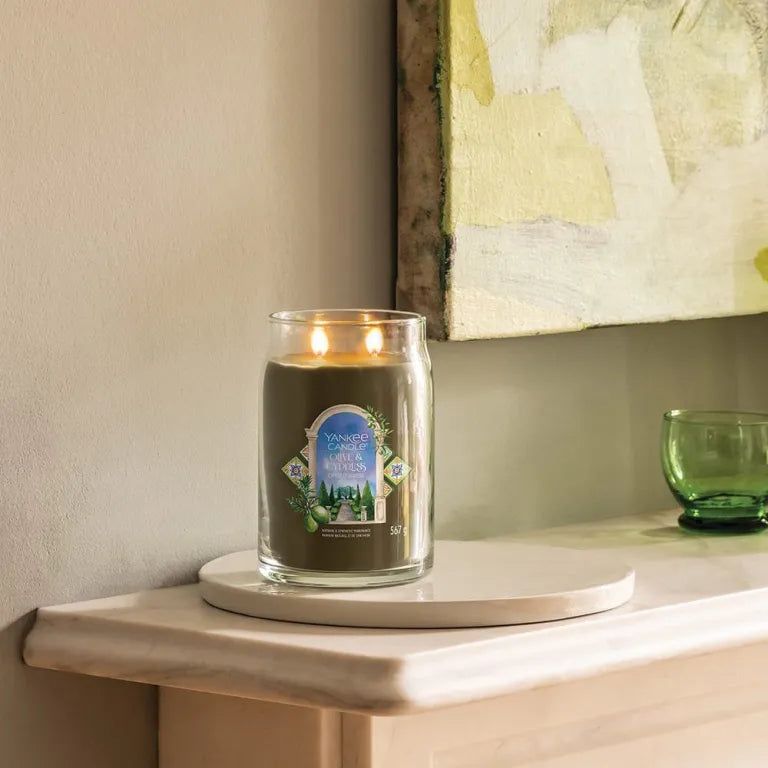 Candela Yankee Candle Olive & Cypress accesa, in vaso di vetro. Su vassoio bianco, davanti a muro con opera d'arte.