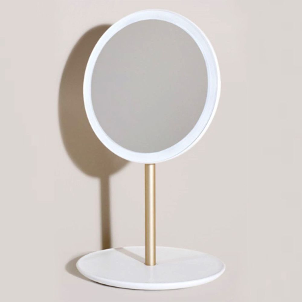 Specchio rotondo a LED su supporto dorato con base bianca, di profilo.